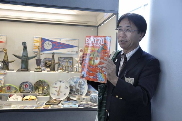 関西ウォーカー編集「EXPO70 グッズコレクション」を手に、展示品の解説をする白井達郎万博ミュージアム館長