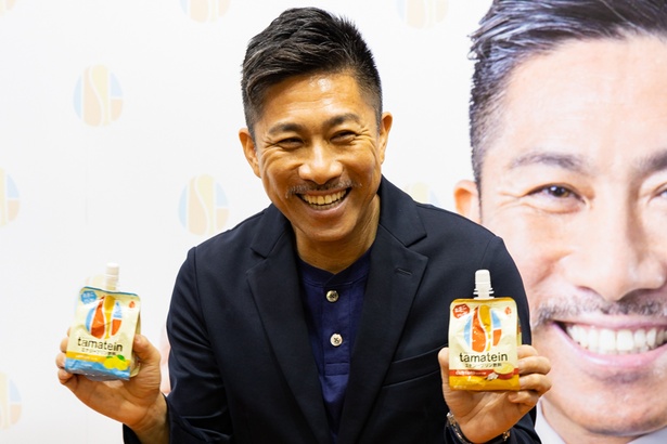 飲むエナジープリン「たまテイン」を持って笑顔の前園さん