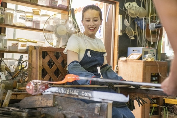 吹きガラス作りに悪戦苦闘しながらも笑顔を見せるドーキンズさん。写真は形成したガラスに柄となるガラスを巻き付けているようす