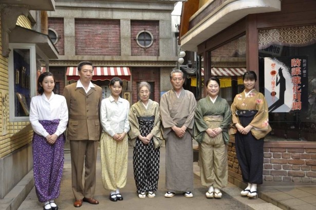 神戸の街のセットに並んだ出演者。張り紙の言葉が時代背景を語る。左から連佛美沙子、生瀬勝久、芳根京子、中村玉緒、本田博太郎、山村紅葉、三倉茉奈